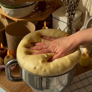 Può includere: Una mano preme su una grande palla di pasta in una ciotola di miscelazione argentata. L'impasto è giallo chiaro e ha una consistenza leggermente ruvida. La ciotola è appoggiata su una superficie di legno. Sullo sfondo ci sono candele e altri oggetti.