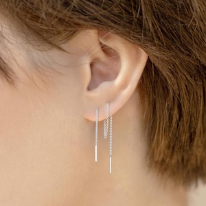 Peut inclure: Une paire de boucles d'oreilles en chaîne argentée avec une petite barre à l'extrémité de chaque chaîne. Les boucles d'oreilles sont portées à l'oreille gauche.