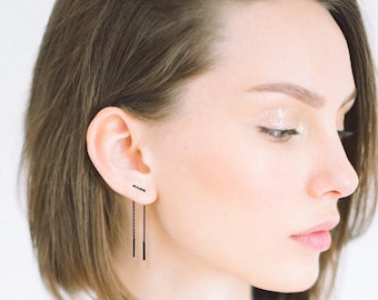 May include: Black linear drop earrings. The earrings feature a small bar stud and a delicate chain that descends to a longer, rectangular bar. The jewelry is minimalist and modern, suitable for everyday wear or special occasions.