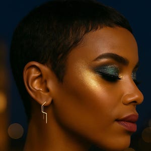 May include: A woman with short dark hair and striking eye makeup, wearing gold geometric earrings. The earrings have a linear, angular design. The background shows blurred city lights.