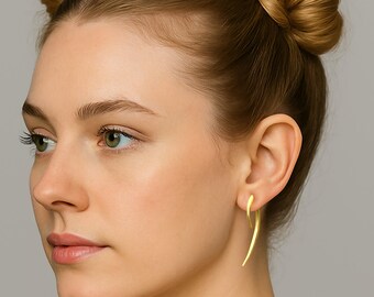 May include: A close-up of a woman wearing gold-colored, curved earrings. The earrings have a smooth, polished finish and a simple, elegant design. The earrings are a bright, shiny gold color.