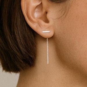 May include: A woman wearing a silver bar earring with a dangling chain.