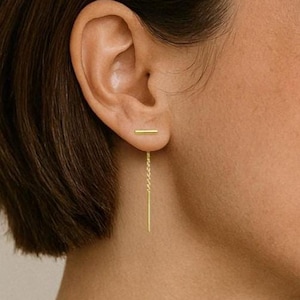 May include: A pair of gold earrings with a thin chain dangling from one earring. The chain is attached to a small bar that is worn in the earlobe.