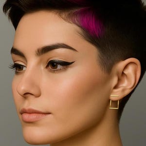 May include: A close-up of a person's face, showcasing a pair of small, square gold earrings. The earrings are a minimalist design, with a square shape. The person has short, dark hair with a streak of pink. The person is wearing black eyeliner.