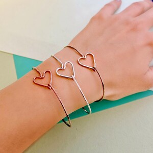 May include: Three silver heart-shaped wire bracelets on a person's wrist. The bracelets are stacked on top of each other.
