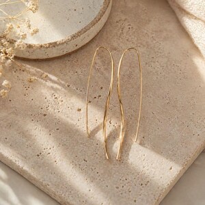 May include: A pair of gold-coloured, elongated drop earrings. The earrings have a curved, open design, with a smooth, polished finish. They are displayed on a textured, light-coloured surface, with a neutral background.