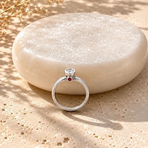 May include: A silver ring featuring a textured band with a clear round stone set at the top and a small red circular accent stone positioned directly below the main setting, creating a distinct contrast against the metallic surface of the band.