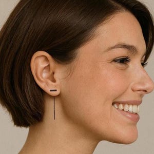 May include: A woman with short black hair wearing a black bar earring with a dangling chain.