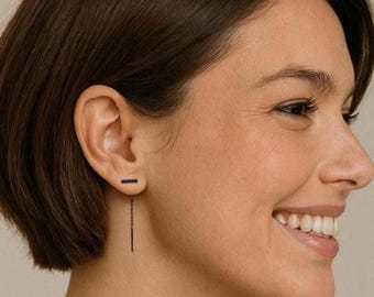 May include: A woman with short black hair wearing a black bar earring with a dangling chain.