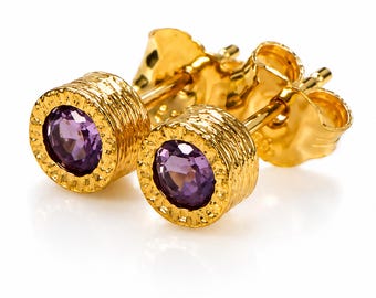 May include: A pair of gold stud earrings featuring a round, faceted purple amethyst gemstone set in a textured, cylindrical gold casing with a notched rim and a standard post back closure, showcasing a polished metallic finish throughout.