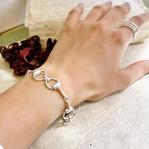 May include: A polished silver chain bracelet featuring a repeating horse bit link design. The bracelet is worn on the wrist, and a matching silver ring with three thin bands is worn on the finger. The metal has a bright, reflective finish.