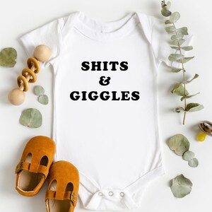 May include: White baby onesie with the text "SHITS & GIGGLES" in black. The onesie is laid flat on a white surface with brown baby shoes and a wooden rattle.