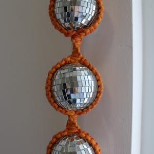 Könnte beinhalten: Eine Schnur mit drei Discokugeln, jede in orangefarbenem Makramee eingefasst, hängt an einer weißen Wand.