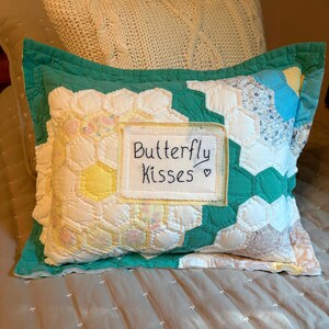 May include: A rectangular throw pillow featuring a patchwork design of white, yellow, and teal hexagonal fabric pieces. A central white fabric label is stitched onto the front with the black embroidered text Butterfly Kisses and a heart.