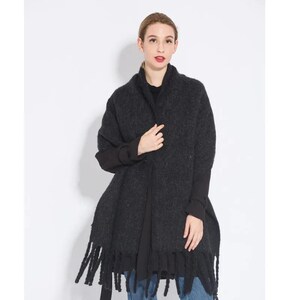 May include: A black fringed scarf made of a soft, fuzzy material. The scarf is draped around the neck of a person wearing a black shirt and blue jeans.