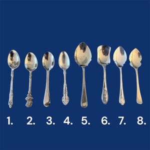 May include: Eight silver spoons of different designs are displayed against a dark blue backdrop. Each spoon has a unique bowl shape and handle detail. Numbers 1 to 8 are below the spoons.