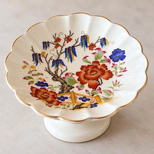 May include: A white ceramic pedestal bowl with a scalloped rim and a gold-painted edge. The interior features a colourful floral pattern with large orange-red flowers, blue hanging blossoms, green leaves, and a small yellow fence illustration.