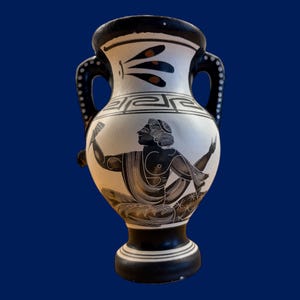 May include: A black and white ceramic amphora with two handles. The vase features a painted scene of a figure in a toga, with geometric patterns and decorative elements. The rim and base are black, and the background is white.