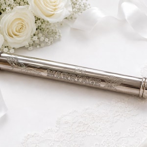 May include: A cylindrical silver-toned metal certificate holder featuring the engraved word MARRIAGE centered on the body, flanked by detailed floral and leaf motifs. The tube has rounded end caps and a polished, reflective metallic finish.