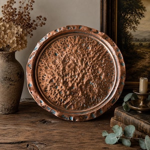 May include: A round copper tray with a hammered texture and a distressed rim. The tray has a reddish-brown hue with areas of blue-green oxidation. The tray is likely vintage or antique, and is displayed on a wooden surface.