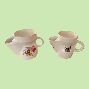 May include: Two vintage shaving mugs. One mug features a floral design with pink, purple, and yellow flowers. The other mug has a black and white dog illustration. Both mugs are white with a spout and handle.