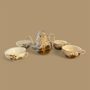 May include: A vintage porcelain tea set featuring a teapot and four teacups. The set is primarily white with gold trim and hand-painted landscape scenes. The teapot has a lid and a curved spout. The teacups have handles.