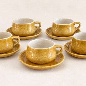 May include: A set of five ceramic tea cups and matching saucers in a mustard yellow glaze with a subtle crackle texture. Each cup features a rounded body, a side handle, and a smooth white interior, arranged in a group on a neutral surface.