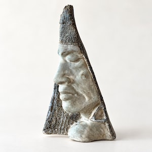 May include: A triangular ceramic sculpture featuring a human face in profile with closed eyes. The piece has a textured, weathered surface with a muted grey and brown color palette, showcasing detailed facial features and a pointed top edge.