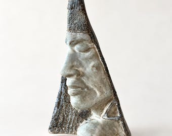 May include: A triangular ceramic sculpture featuring a human face in profile with closed eyes. The piece has a textured, weathered surface with a muted grey and brown color palette, showcasing detailed facial features and a pointed top edge.