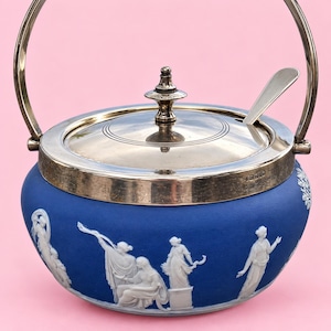 May include: A blue ceramic sugar bowl with white relief figures, featuring a silver-plated metal lid, a matching swing handle, and a small silver spoon. The lid has a central finial and the body displays classical figures in white bas-relief.