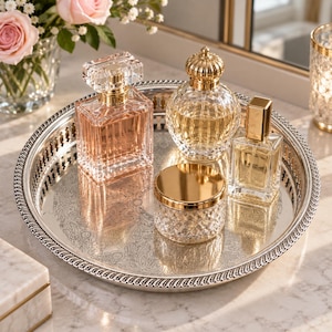 May include: A round silver-toned metal tray with an ornate engraved pattern and a decorative rim, holding three glass perfume bottles and a small glass jar with a gold-toned lid. The bottles contain liquids in shades of pink and gold.