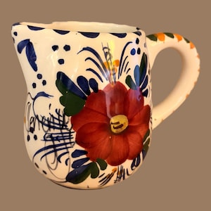 May include: White ceramic pitcher with a large red flower and green leaves. The pitcher is decorated with blue and white floral patterns and has an orange and green handle. A decorative piece for the kitchen or dining room.