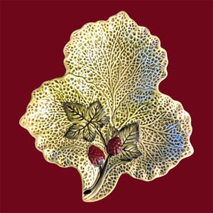 May include: A ceramic leaf-shaped dish with a textured, pale green surface. The dish features a branch with two red berries and green leaves. The background is a deep red.