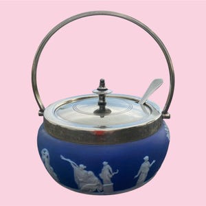 May include: A blue and silver sugar bowl with a handle and spoon. The bowl has a silver lid and rim, and a silver handle. The blue body of the bowl features white relief figures. The spoon rests on the rim.