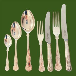 May include: A set of silver-plated cutlery, comprising spoons, forks, and knives, displayed against a green backdrop. The spoons are of different sizes, and the knives are marked "Arthur Price of England". The handles have an elaborate pattern.