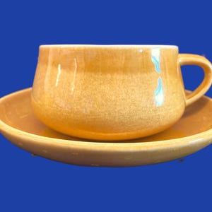 May include: A yellow ceramic teacup and saucer set. The cup has a rounded shape and a handle. The saucer has a slightly scalloped edge.