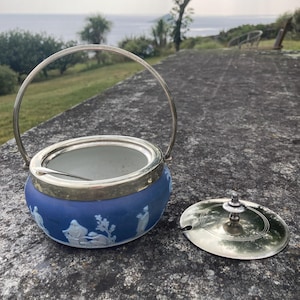 May include: A blue and white ceramic sugar bowl with a silver rim and handle. The bowl features a classical relief design and comes with a silver lid and spoon. The bowl is sitting on a stone surface.