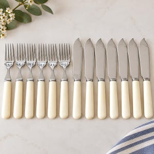 May include: A set of six silver-toned forks and seven silver-toned knives arranged in two rows. Each piece of cutlery features a smooth, cream-colored handle with a rounded end. The metal heads of the forks have four tines, while the knives have a curved, flat blade design.