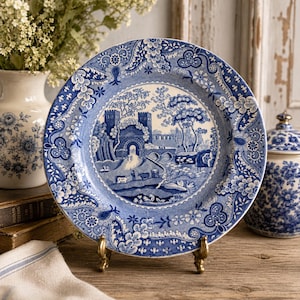 May include: A round ceramic plate featuring a blue and white transferware pattern. The center depicts a landscape with a stone castle, a bridge, and figures. The wide rim is decorated with intricate floral and foliate motifs in shades of blue.