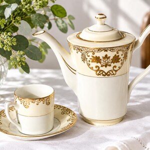 May include: A cream-coloured porcelain teapot and a matching teacup with a saucer, both featuring intricate gold filigree patterns and floral motifs along the upper rims. The teapot has a tall, cylindrical body, a curved spout, and a lid with a gold knob.