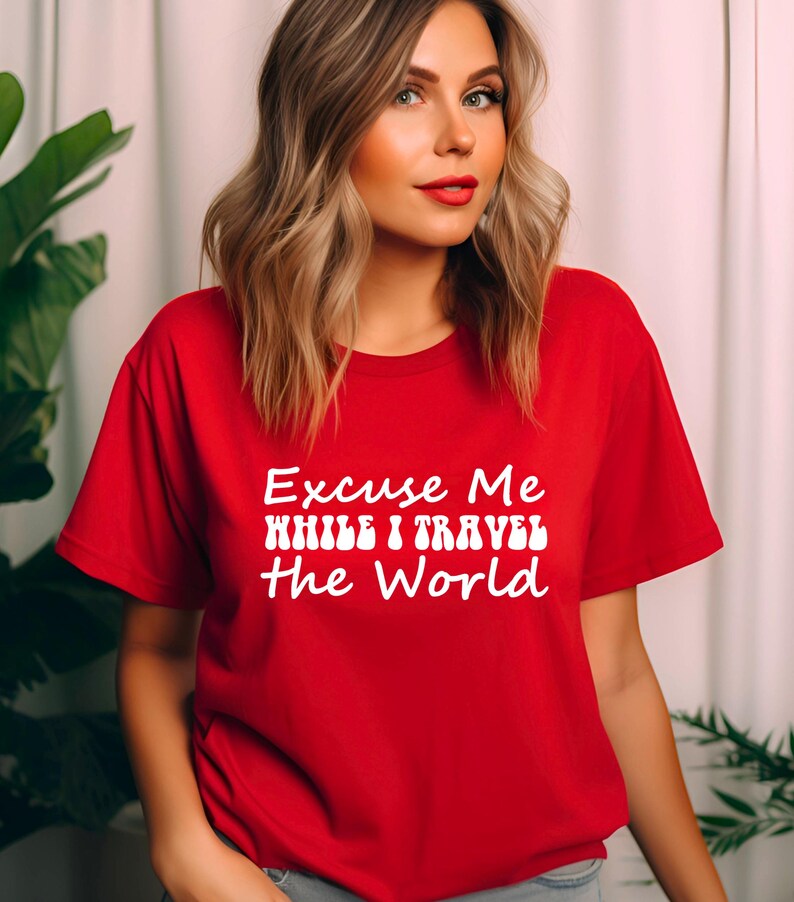 May include: A red t-shirt featuring the white text "Excuse Me While I Travel The World". The shirt has a crew neck and is likely made from a soft fabric. The design suggests a theme of travel and exploration.