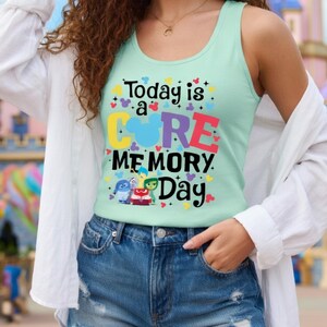 May include: A light green tank top with the text "Today is a Core Memory Day" in colorful letters. The shirt features cartoon characters and heart shapes. The person is wearing denim shorts and a white shirt.