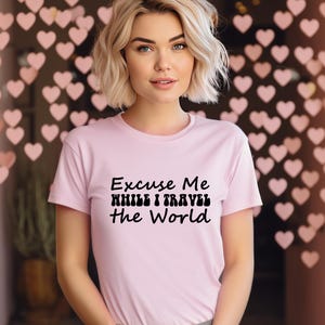 May include: A pale pink t-shirt featuring the black text "Excuse Me While I Travel The World". The t-shirt has a crew neck and is worn by a person with blonde hair.