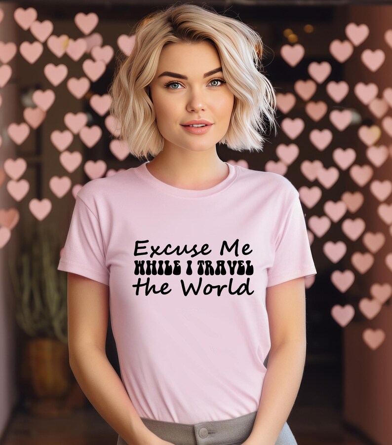 May include: A pale pink t-shirt featuring the black text "Excuse Me While I Travel The World". The t-shirt has a crew neck and is worn by a person with blonde hair.