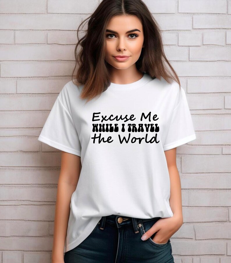 May include: White t-shirt with the text "Excuse Me While I Travel The World" in black. The shirt has short sleeves and a crew neck. The person is wearing dark blue jeans.