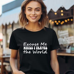 May include: Black t-shirt with the white text "Excuse Me While I Travel The World". The shirt is short-sleeved and has a crew neck. The model is smiling and the background is blurred.