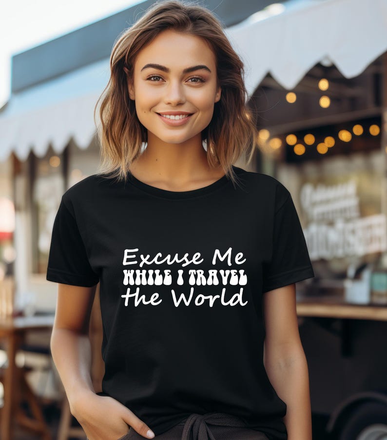 May include: Black t-shirt with the white text "Excuse Me While I Travel The World". The shirt is short-sleeved and has a crew neck. The model is smiling and the background is blurred.