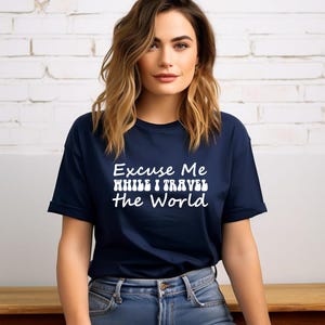 May include: A navy blue t-shirt featuring the white text "Excuse Me While I Travel The World". The shirt has a crew neck and short sleeves. The model is wearing blue jeans.