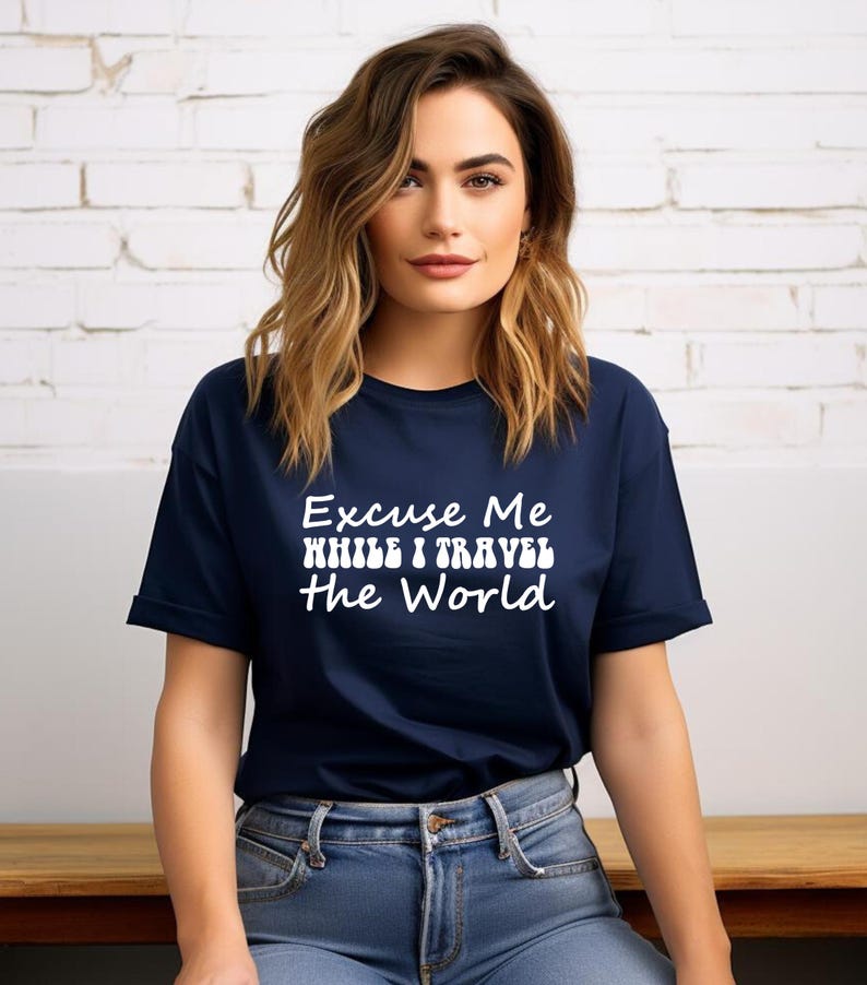 May include: A navy blue t-shirt featuring the white text "Excuse Me While I Travel The World". The shirt has a crew neck and short sleeves. The model is wearing blue jeans.