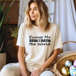 May include: A cream-coloured t-shirt with the text "Excuse Me While I Travel The World" in black. The shirt is paired with light-coloured trousers. A wicker basket with decorative Easter eggs is in the foreground.